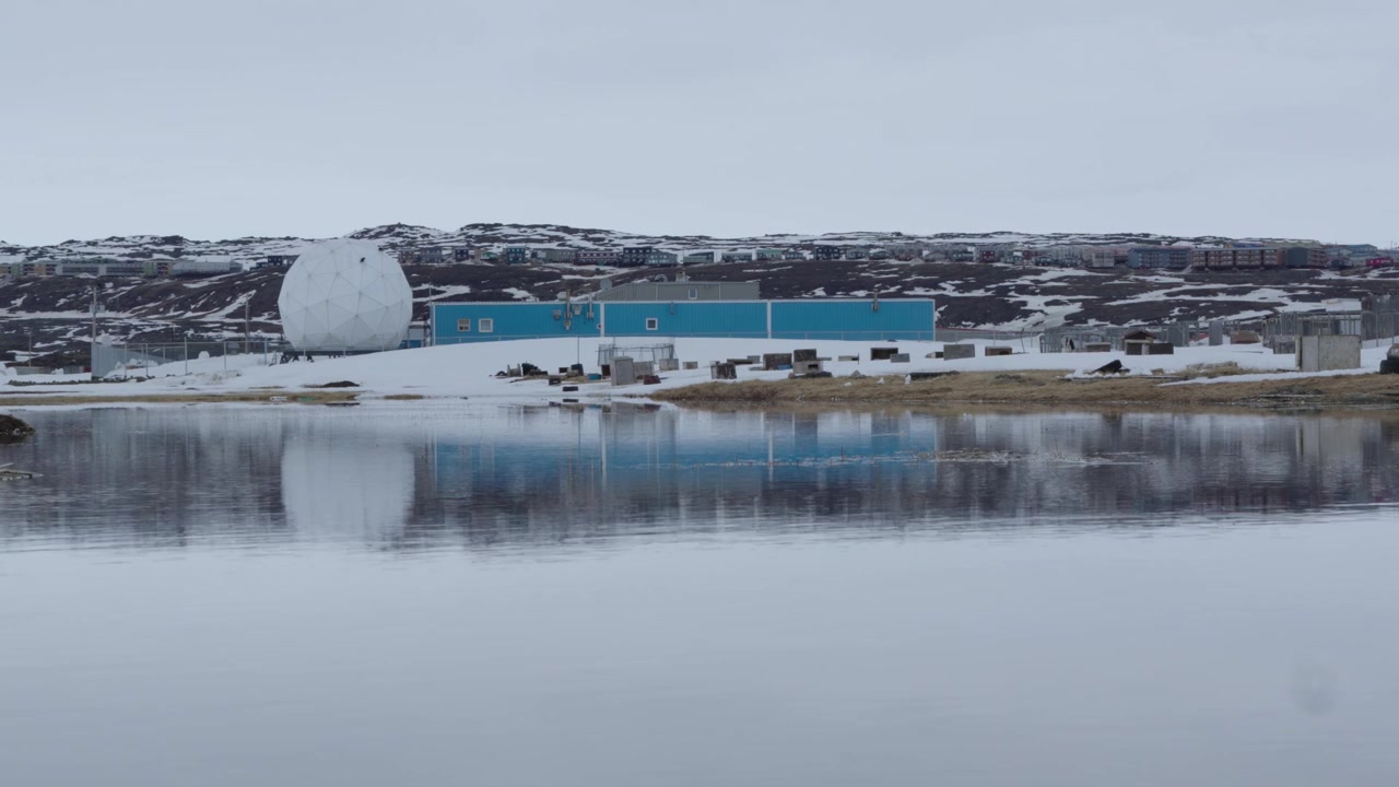 Kamerabilde fra bakkenivå av en stor hvit radarkuppel og en blå industribygning perfekt speilet i et rolig vannbasseng under vårløsningen i Iqaluit.