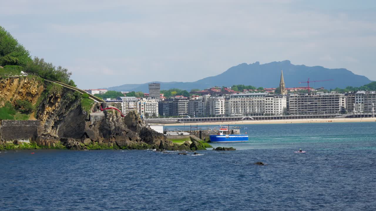 Blue and white boat docked near rocky cliffs with the city skyline in the distance