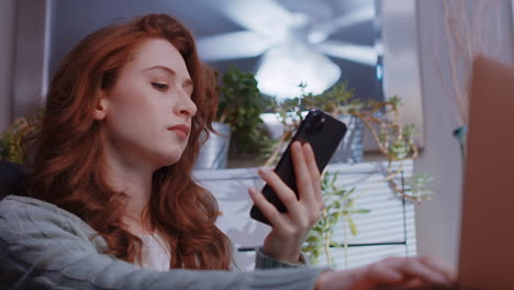 a young caucasian woman is working from home on her laptop and looking at her cell phone