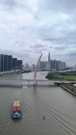 container boats on saigon river, ho chi minh city, with aerial view of ba son suspension bridge with landmark building in vertical format as boat pass along river