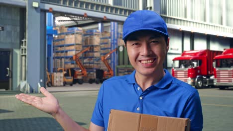 close up of asian male courier in blue uniform smiling and pointing to the side while delivering a carton, outside of logistics distributions warehouse