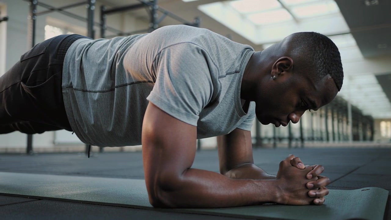 Bracing forearms, man wearing gray shirt, dark shorts holding plank on mat in gym building core