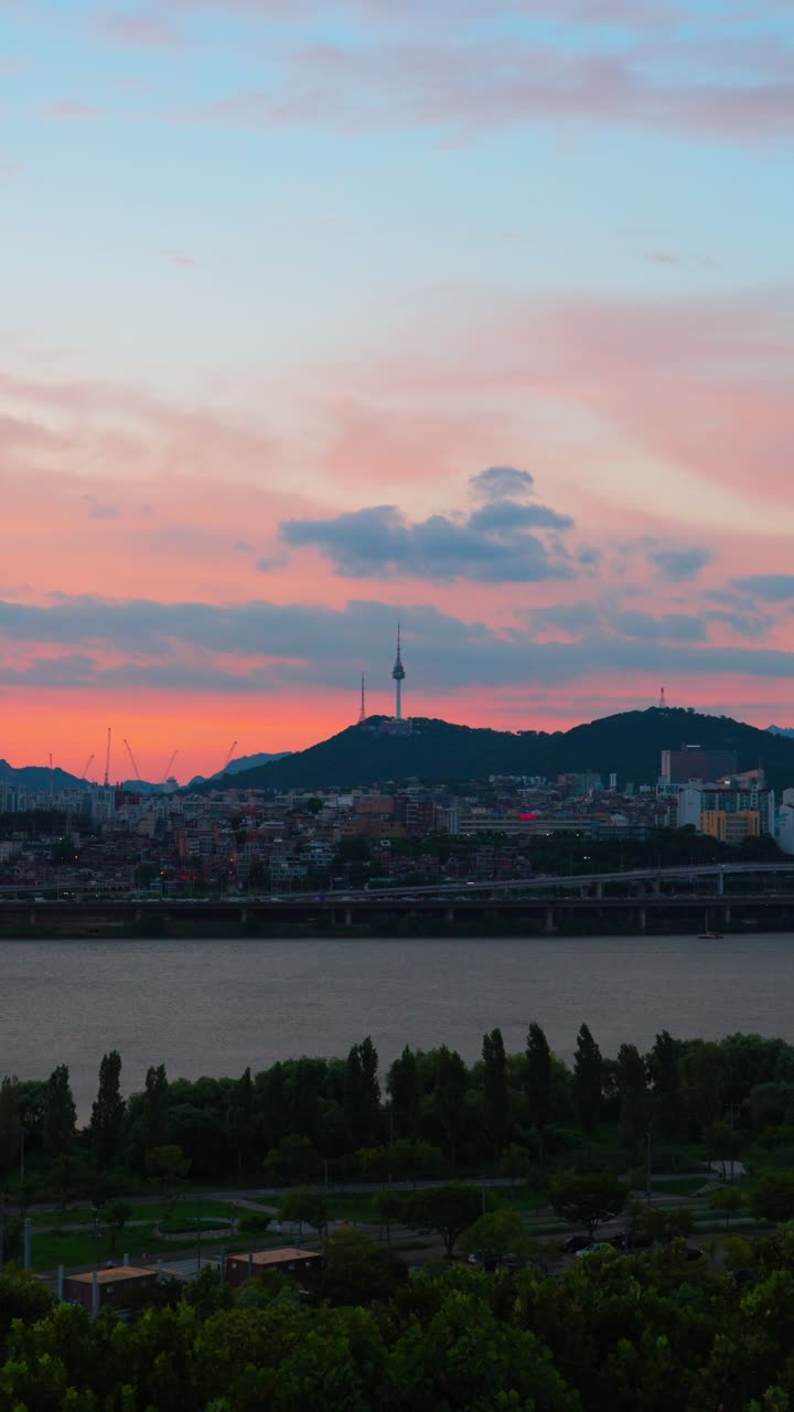 Soaring vertical aerial shot of Hangang Park and Han River, cityscape with N Seoul Tower, all under a calm sunset sky glowing with gentle pastel pink and blue tones