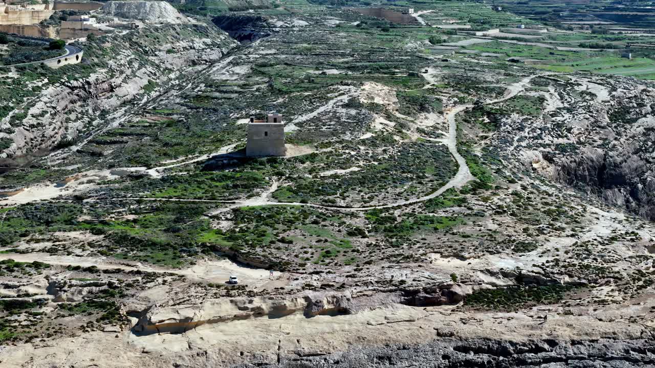 Prospettiva aerea della storica Torre di Dwejra in mezzo al frastagliato paesaggio costiero calcareo dell'isola di Gozo, Malta, durante una luminosa giornata di sole primaverile, con tonalità terrose e vegetazione mediterranea.