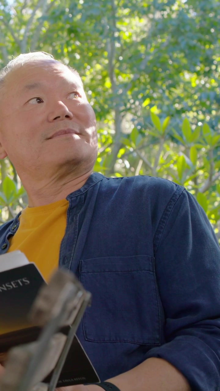 Video vertical: Hombre asiático con camiseta amarilla sosteniendo un libro, mirando fuera de cámara y retomando la lectura en un parque.