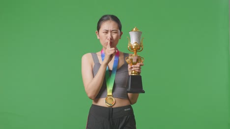 asian woman with a gold medal and trophy making shh gesture telling it is a secret to win as the first winner on green screen background in the studio