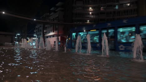 A-blue-tram-passes-by-the-small-fountain-heading-towards-the-shopping-mall-at-night,-Montpellier---France