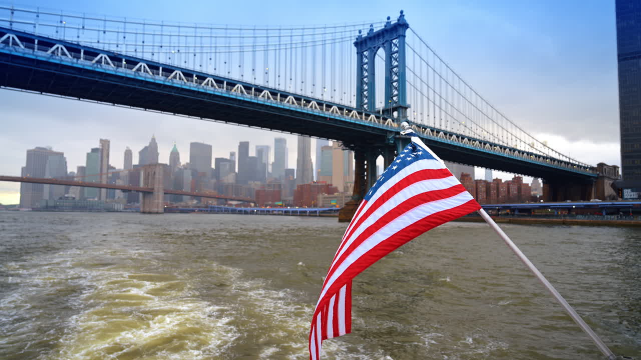 Bandiera statunitense che sventola vicino al Ponte di Manhattan sull'East River. Bandiera americana in primo piano con il ponte sospeso blu e lo skyline della città sullo sfondo.