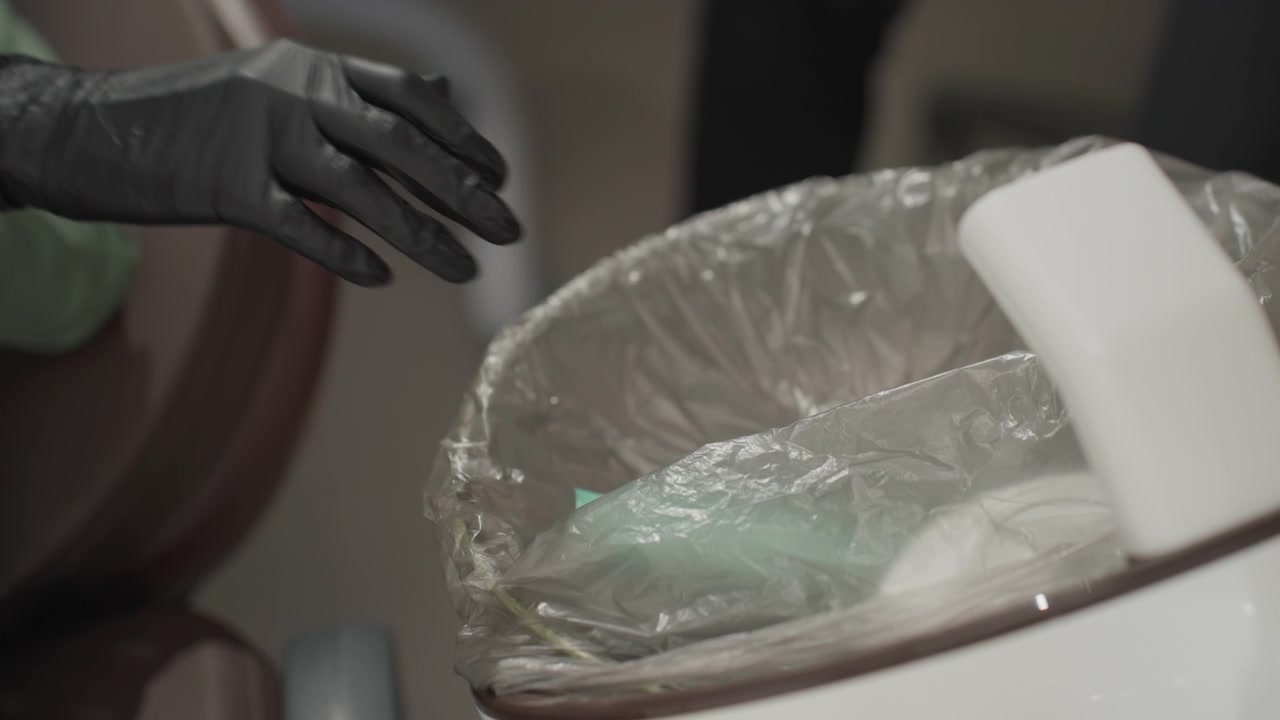 Primo piano di un'assistente dentale che indossa guanti neri e getta guanti verdi usati in un cestino rivestito di plastica durante una procedura ortodontica. La scena si concentra sul movimento della mano guantata e sui rifiuti.