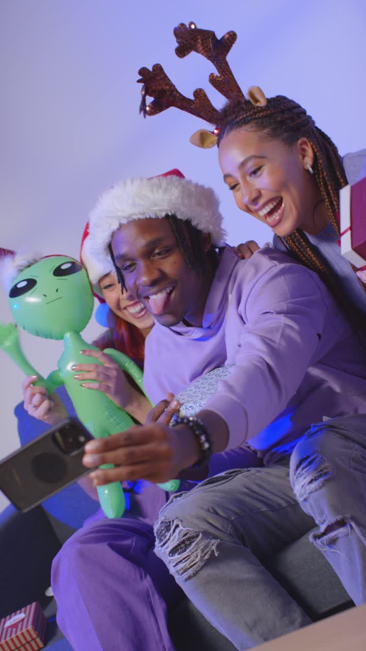 video de estudio vertical de amigos de la generación z en navidad sentados en el sofá con sombrero de papá noel y cuernos de renos tomando selfies en el teléfono móvil 2