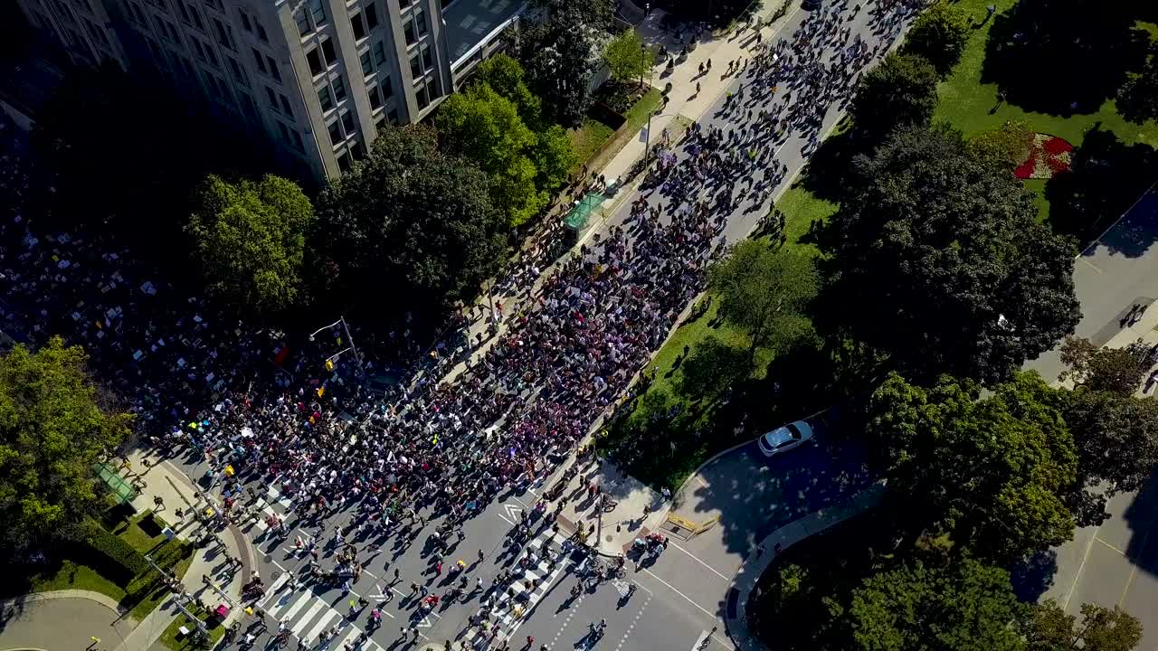 Large group of people flows through downtown Toronto, aerial drone pan. Lots of protesters with democratic free speech signs walk on city roads together, wide daytime exterior in 4k