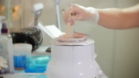 una esteticista se prepara para depilarse las piernas aplicando cera a una espátula de una olla xax