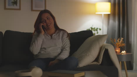 Primer-Plano-De-Una-Mujer-Sonriente-Sentada-En-Un-Sofá-En-Casa-Por-La-Noche-Hablando-Por-Teléfono-Móvil-Filmada-En-Tiempo-Real