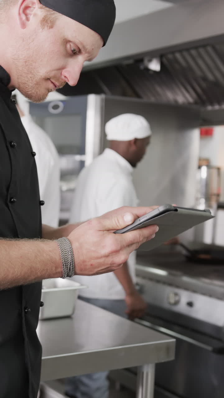 Focused caucasian male chef using tablet in kitchen, slow motion, vertical