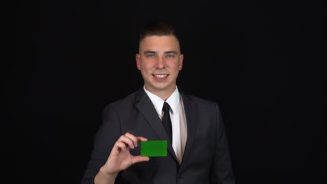 a young businessman in a suit holds a bank card in hand. isolated black background. chromakey green card.