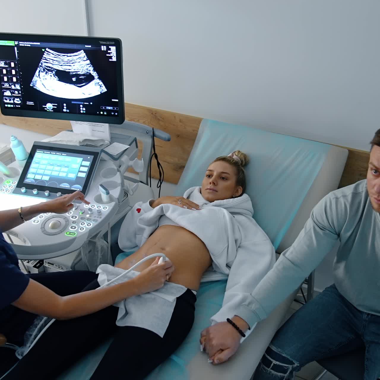 Future parents are in the doctor's cabinet at pregnancy check up. Caucasian lady lies on the couch undergoing ultrasound examination. Worried man holding wife's hand. Top view
