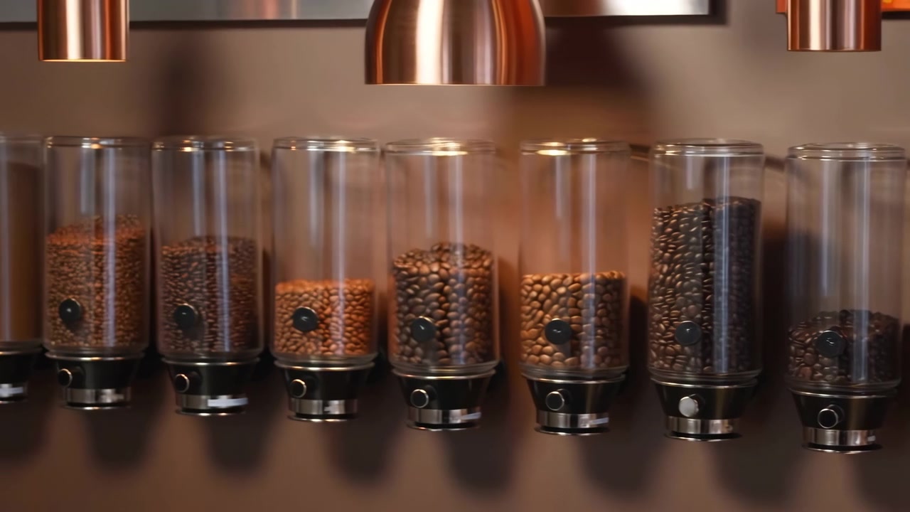 Tight horizontal view of clear cylindrical coffee dispensers mounted on brown wall, filled with roasted beans at different levels, with warm reflections, metallic accents, and clean retail cafe