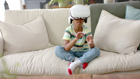 un niño afroamericano con auriculares de realidad virtual se sienta con las piernas cruzadas en un sofá en casa
