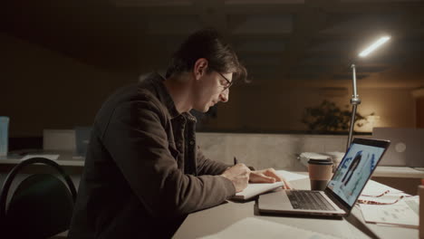 man video calling with colleague and taking notes in office at night