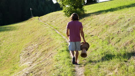 young man with red tshirt, long brown hair and drum in his hand is walking on a small path on a beautiful meadow