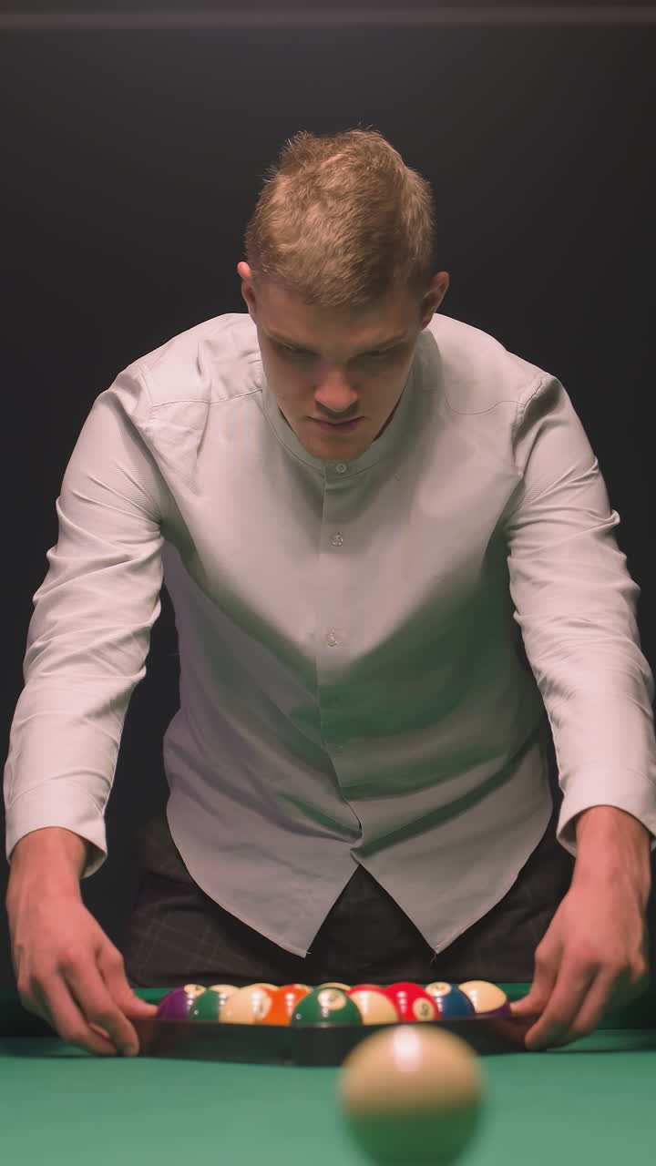 primer plano de un joven en camisa intensamente enfocado en la mesa de billar mientras se prepara para romper. sus manos se mueven sobre un estante de bolas de colores en fieltro verde vibrante, luz suave contra un fondo oscuro
