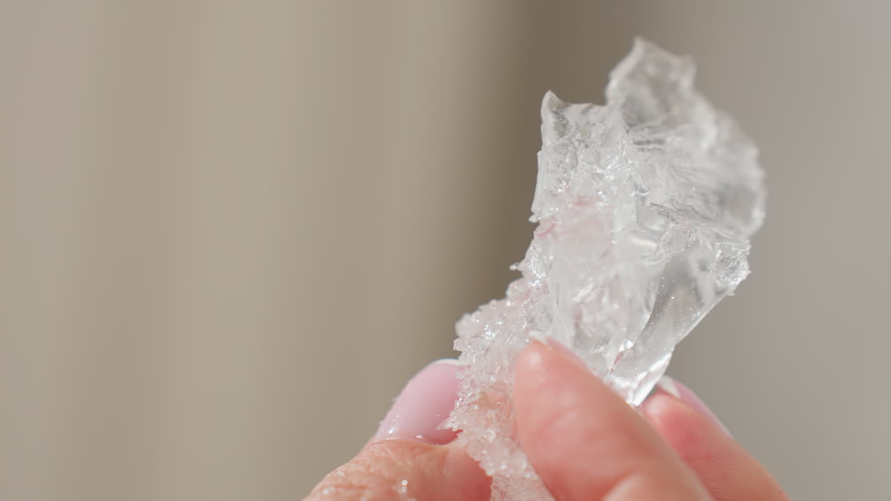 Close up of delicate hand with pink manicured nails gently shaking transparent rubber-like soluble material, highlighting intricate texture and clarity, set against soft neutral background