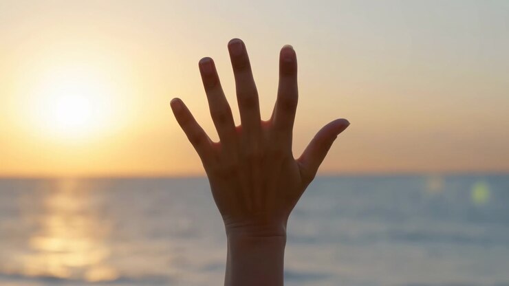 Een open hand blijft geheven voor de zee terwijl de gouden zon vanaf de horizon schijnt, met zachte luchtkleuren, een reflecterend wateroppervlak en een gecentreerd close-up standpunt dat een stil buitenmoment toont.