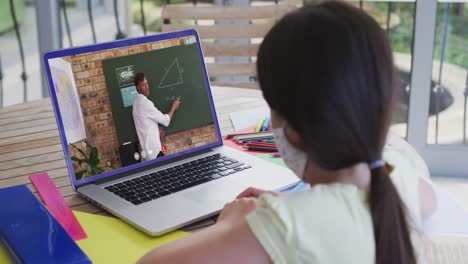 Caucasian-schoolgirl-in-face-mask-using-laptop-on-video-call-with-male-teacher