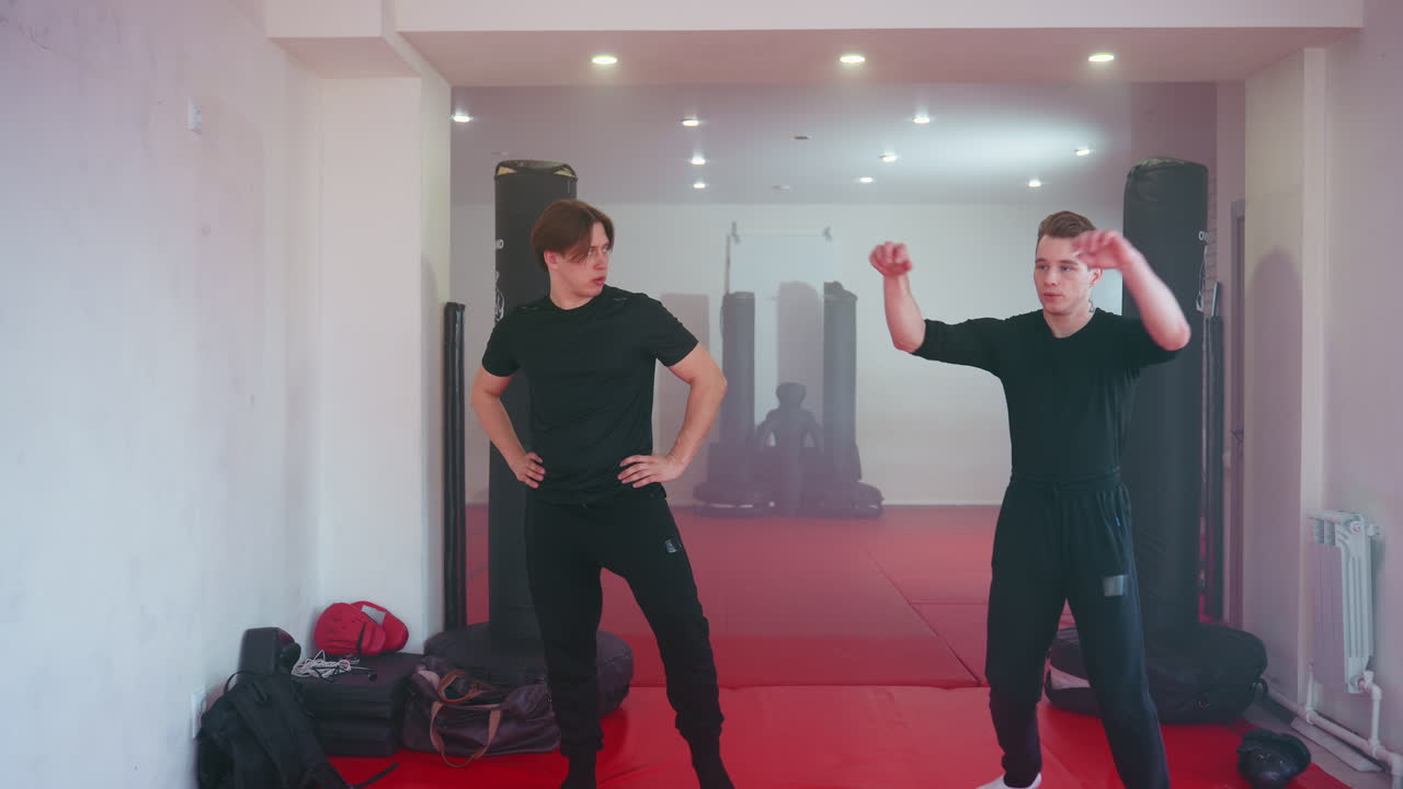 Two fighters in black sportswear prepare on red training mat inside martial arts gym, standing near punching bags with determined stances, focusing on combat training and practicing readiness