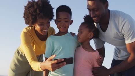 Smiling-african-american-family-using-smartphone-and-embracing-on-sunny-beach