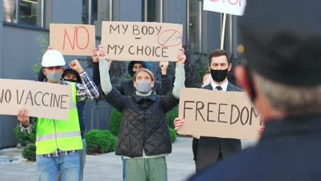 rear view of policeman looking a group of people holding signboards and yelling in a demonstration against covid 19 in the street