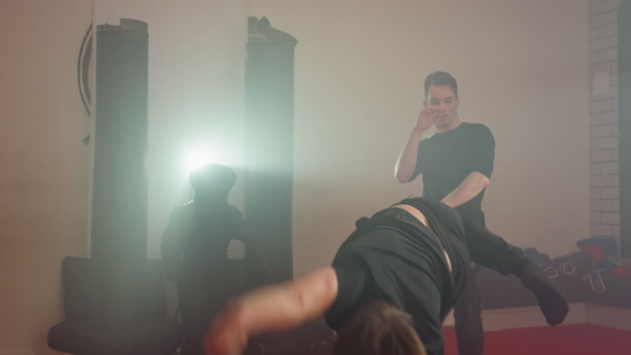 Karate practitioner training in gym holding shield during sparring session with partner, intense practice on red mats, punching bags in background, strong spotlight glare creating dramatic atmosphere