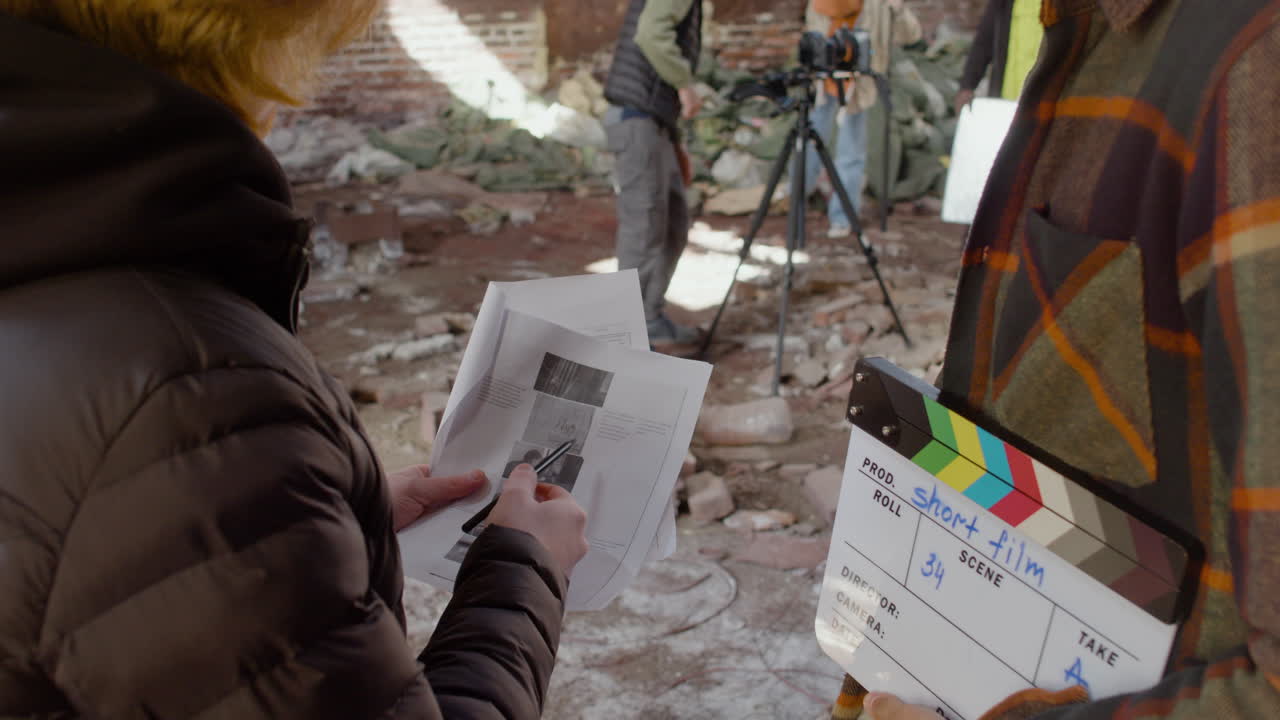 vista de cerca de dos compañeros de producción irreconocibles hablando y leyendo un documento sobre la película en un edificio en ruinas mientras otros colegas se preparan para la grabación