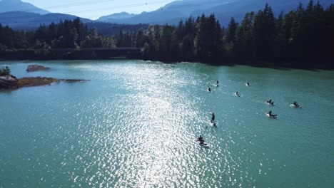 group of stand up paddle boarders, floating in an emerald lake next to a highway surrounded by forest trees, islands mountains on sunny day near whistler british columbia