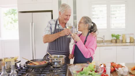 feliz pareja de ancianos diversos usando delantales y bebiendo vino en la cocina