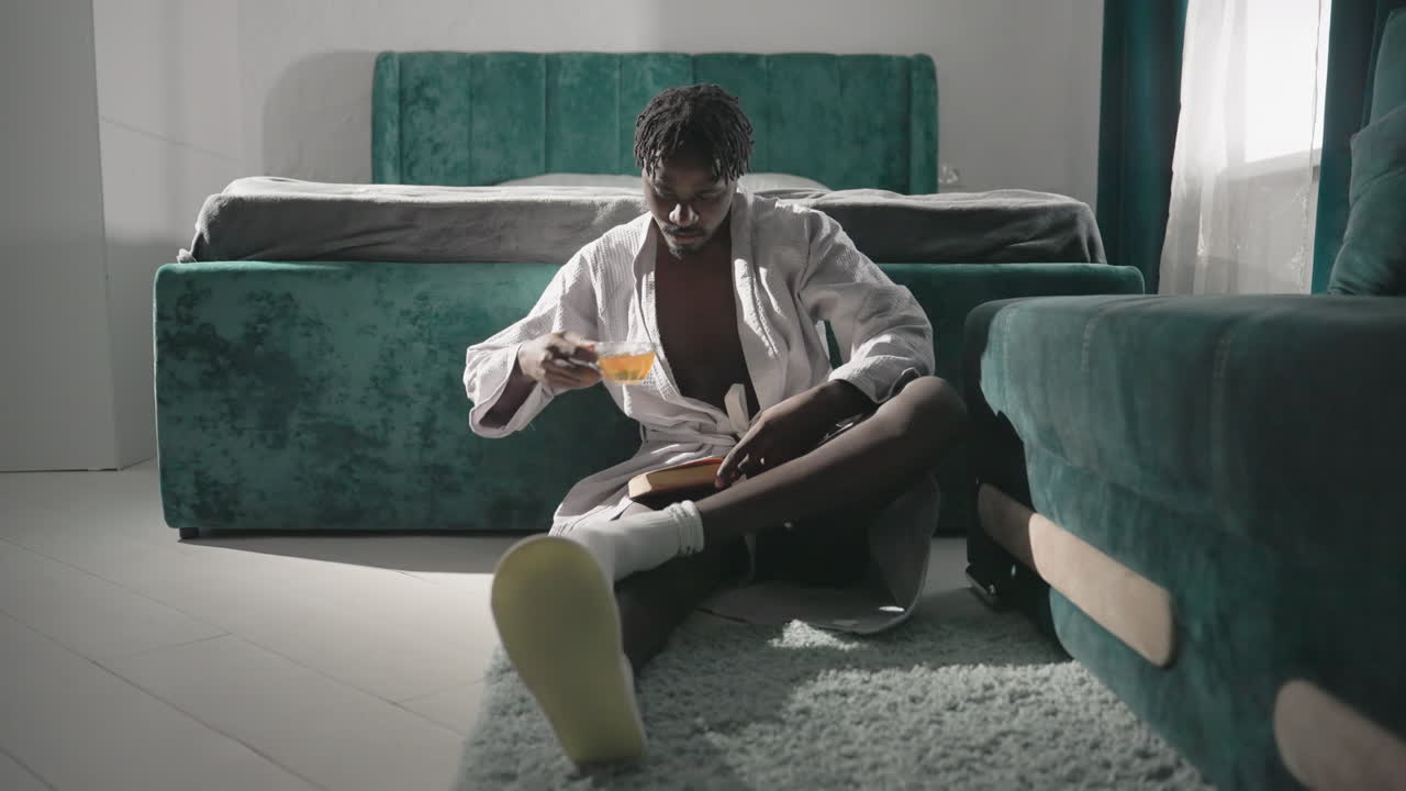 Man sitting on rug in robe, reading book, sipping tea in modern cozy bedroom. Peaceful atmosphere with green furniture, morning light casting soft shadows. Relaxing morning routine in stylish setting