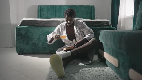 man sitting on rug in robe, reading book, sipping tea in modern cozy bedroom. peaceful atmosphere with green furniture, morning light casting soft shadows. relaxing morning routine in stylish setting