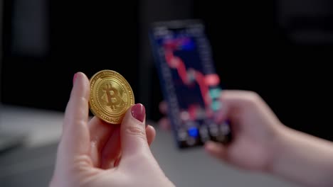 Dedos-Femeninos-Sosteniendo-Bitcoins-Dorados-Viendo-El-Declive-Del-Gráfico-De-Inversión-En-Criptocomercio-En-El-Teléfono-Inteligente