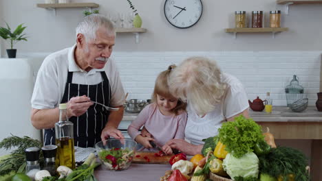 Senior-couple-in-kitchen-teaching-granddaughter-child-how-to-cook,-chopping-pepper-with-knife