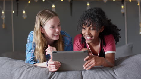 amigas adolescentes felices y diversas acostadas en la cama y usando la tableta en casa, en cámara lenta