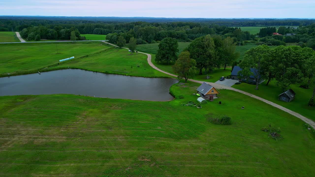 Aerial View of a Lake with Wooden House Surrounded by Grass Fields and Woodland in Latvia