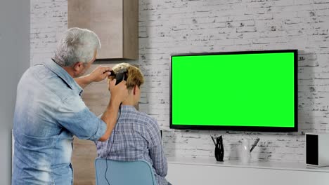 un hombre le corta el cabello a su esposa en casa mientras ve la televisión de pantalla verde