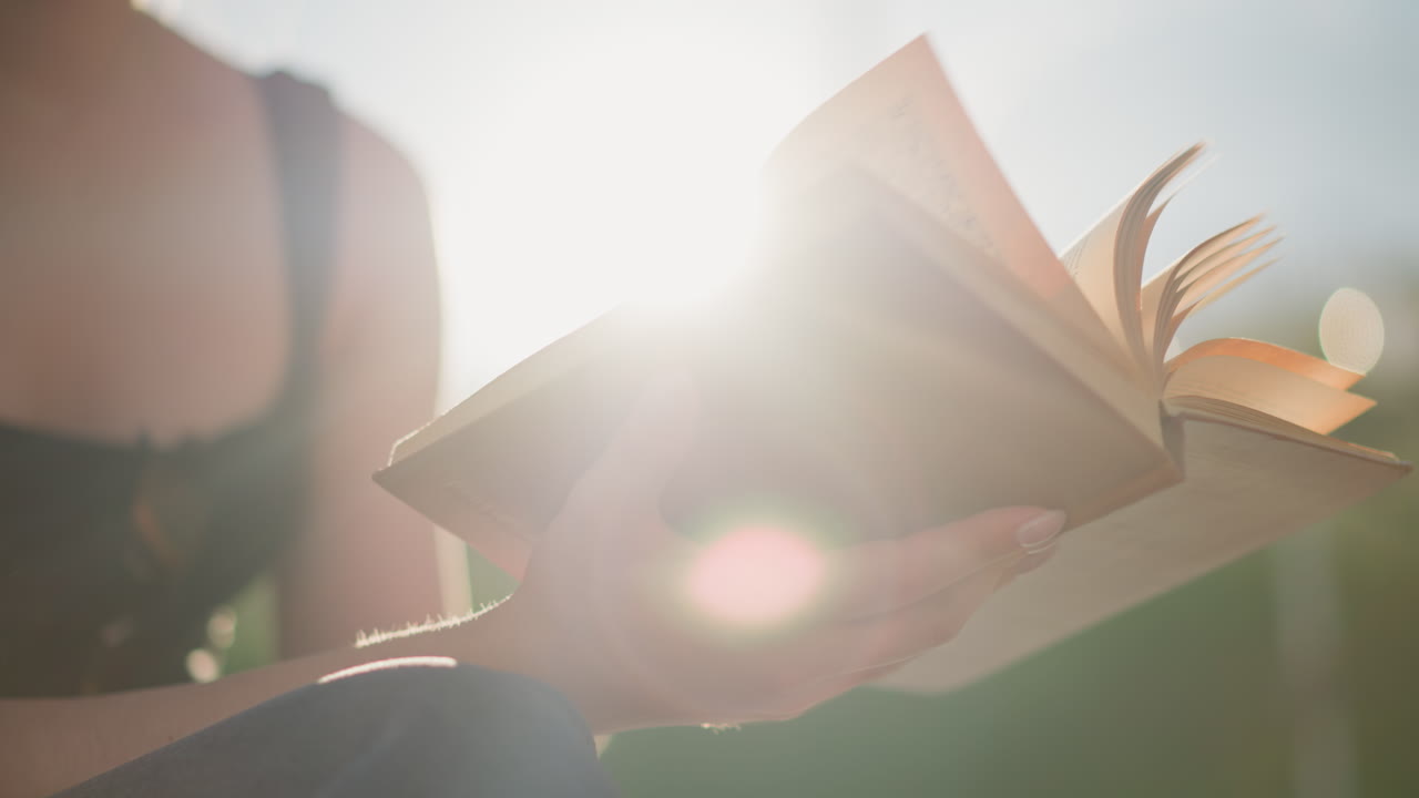 태양 광선은 밖에서 다리를 고 앉아있는 여성이 들고 있는 열린 책에 반사되며, 그녀의 모습은 배경에서 약간 흐릿해지고, 바람은 부드럽게 페이지를 통해 불고 있습니다.