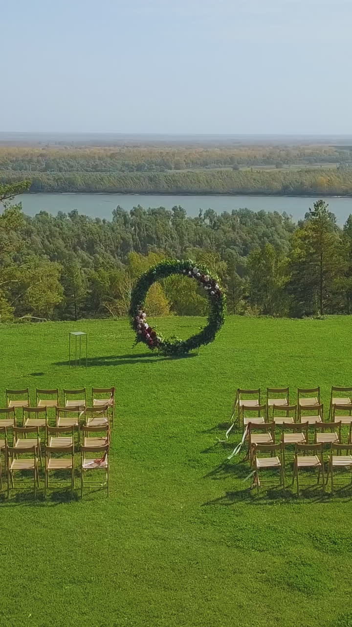 large green plant garland of wedding venue and empty chairs on lush grass on sunny day against wide river upper view