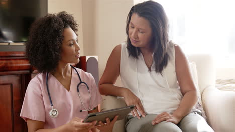 nurse in scrubs holding tablet, discussing health with woman on couch