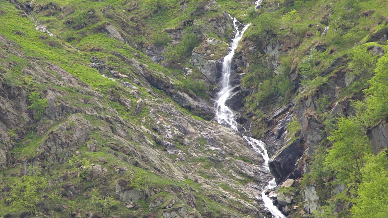 Un'inquadratura ampia cattura una stretta cascata che scorre lungo una scogliera montuosa aspra e verde.
