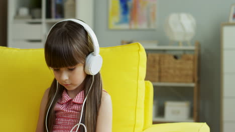 Niña-Bonita-Con-Auriculares-Grandes-Sentada-En-Un-Sofá-Amarillo-En-La-Sala-De-Estar-En-Casa