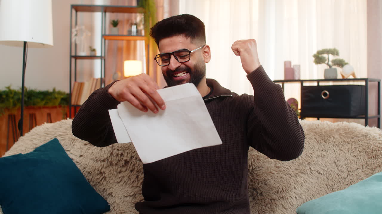 Indian man on sofa opens envelope and celebrates good news after reading positive letter at home