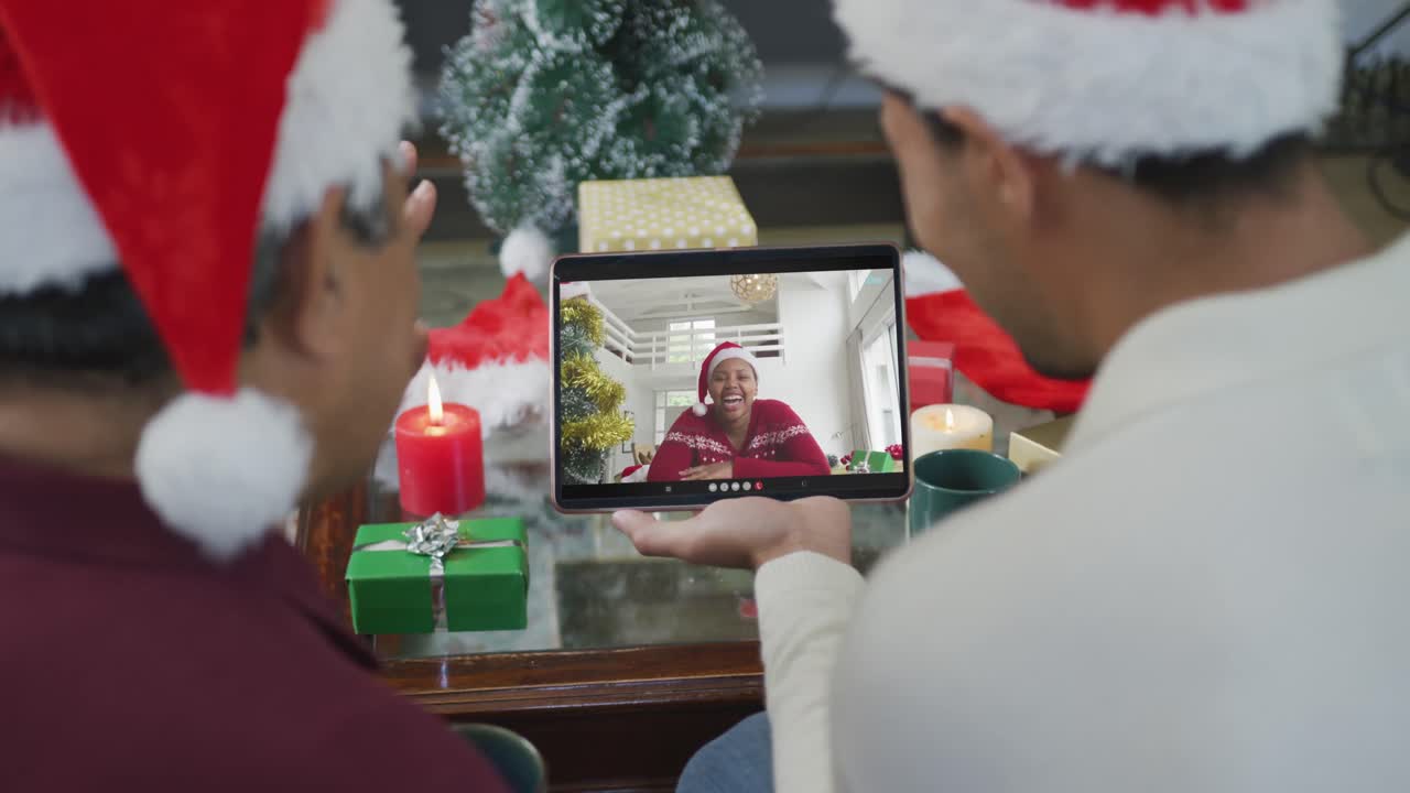 padre y hijo biraciales con sombreros de santa usando una tableta para una videollamada de navidad con una mujer en la pantalla