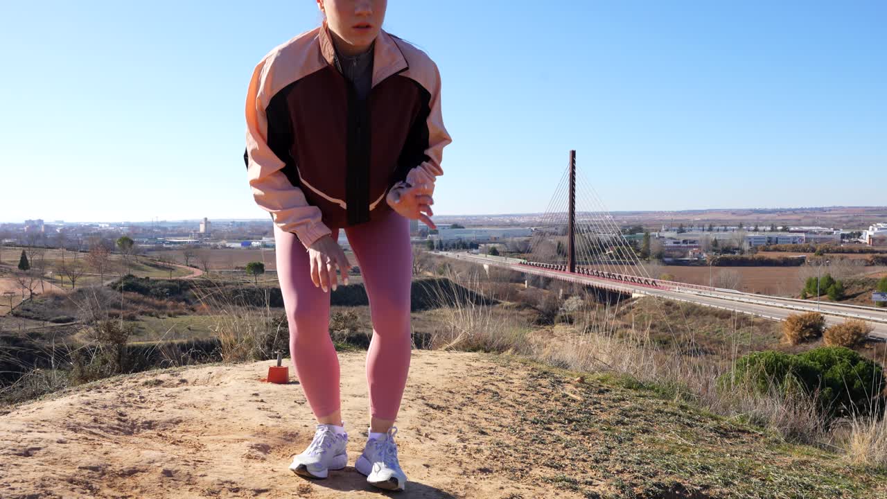 Young woman runs uphill outside city, crouches exhausted and unzips jacket